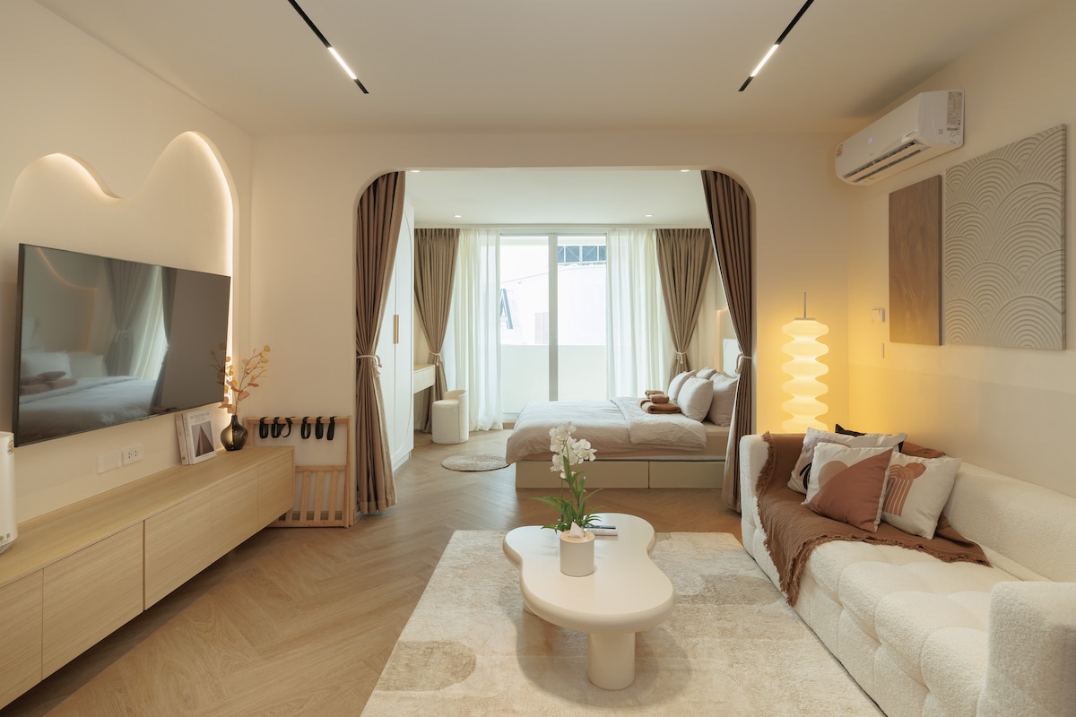 A modern studio layout is displayed, featuring a spacious living area with a light-colored sofa and a soft area rug. A large TV is mounted on the wall, while natural light fills the room through curtain-draped windows. The bedroom area, separated by curtains, features a comfortable queen bed with neutral-toned bedding.