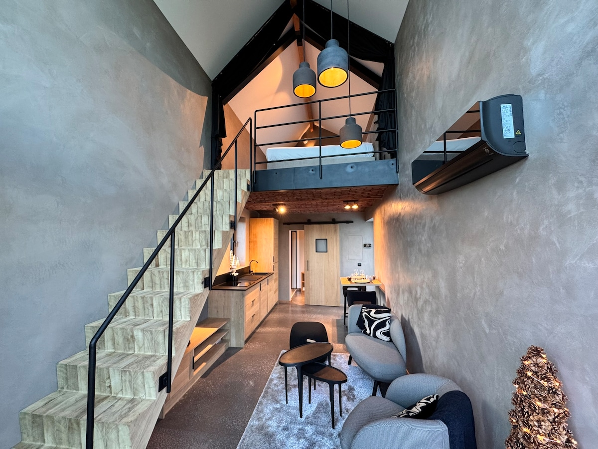 A modern interior showcases an open space with high ceilings and large windows. A stylish staircase leads to a lofted area featuring a king-size bed. Below, a kitchenette and seating area are visible, complemented by soft lighting and a cozy atmosphere.
