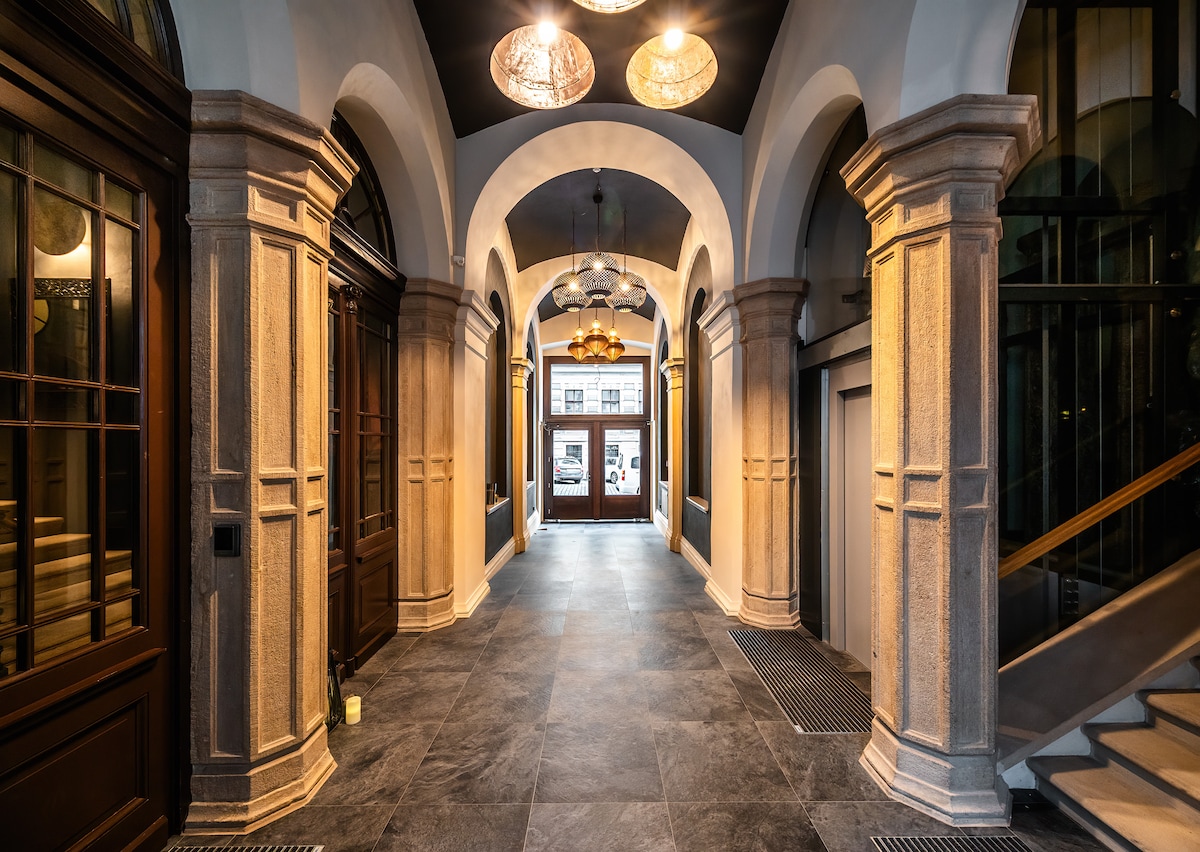 A spacious hallway is framed by elegant archways and complemented by large wooden doors. Soft lighting is provided by decorative fixtures, highlighting the stone columns and dark tile flooring. The area leads to a welcoming entrance, creating an inviting transition into the apartment.