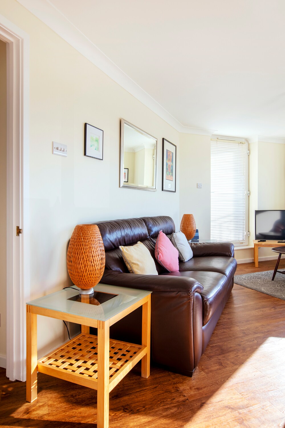 A comfortable living area features a dark leather sofa adorned with colorful cushions. A wooden coffee table is positioned next to the sofa, topped with a decorative lamp. Natural light floods the space through a nearby window, enhancing the inviting atmosphere.