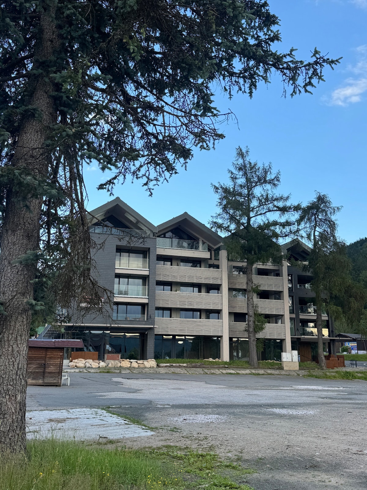A modern four-story building with a triangular roof design is surrounded by tall trees. Large windows adorn the facade, offering a glimpse of the interior. The landscape features a gravel pathway and patches of grass in the foreground, creating a natural setting.