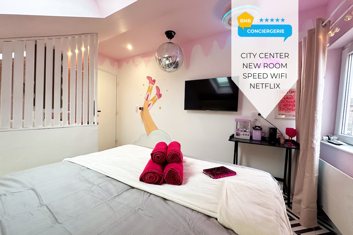 A bright room features a spacious bed adorned with rolled towels in vibrant pink. A disco ball hangs from the ceiling, reflecting light onto the chic pink walls. A modern television is mounted on one wall, and a coffee machine is positioned nearby.