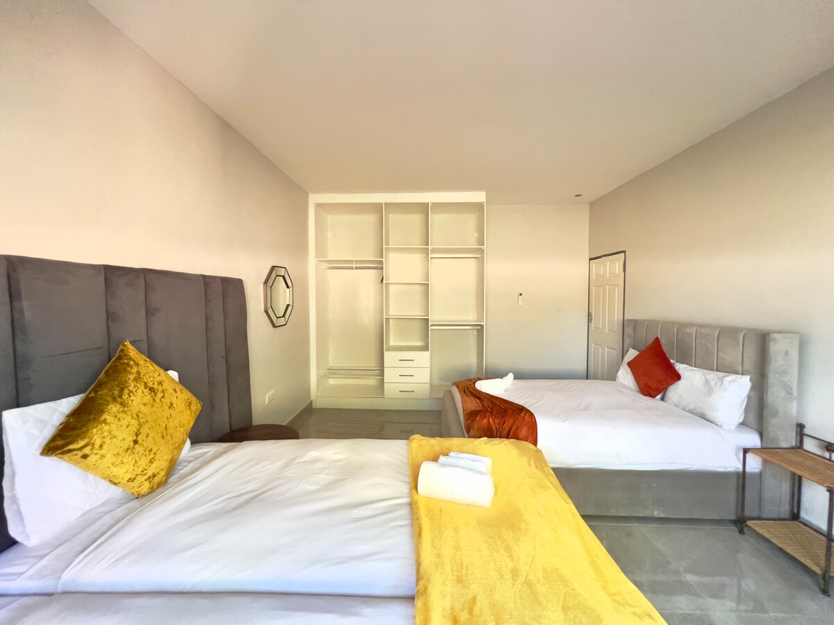 A light-filled bedroom features two spacious beds topped with neutral linens and colorful throw pillows. A wooden side table holds neatly folded towels. A large, empty closet with open shelving is visible, offering ample storage space.