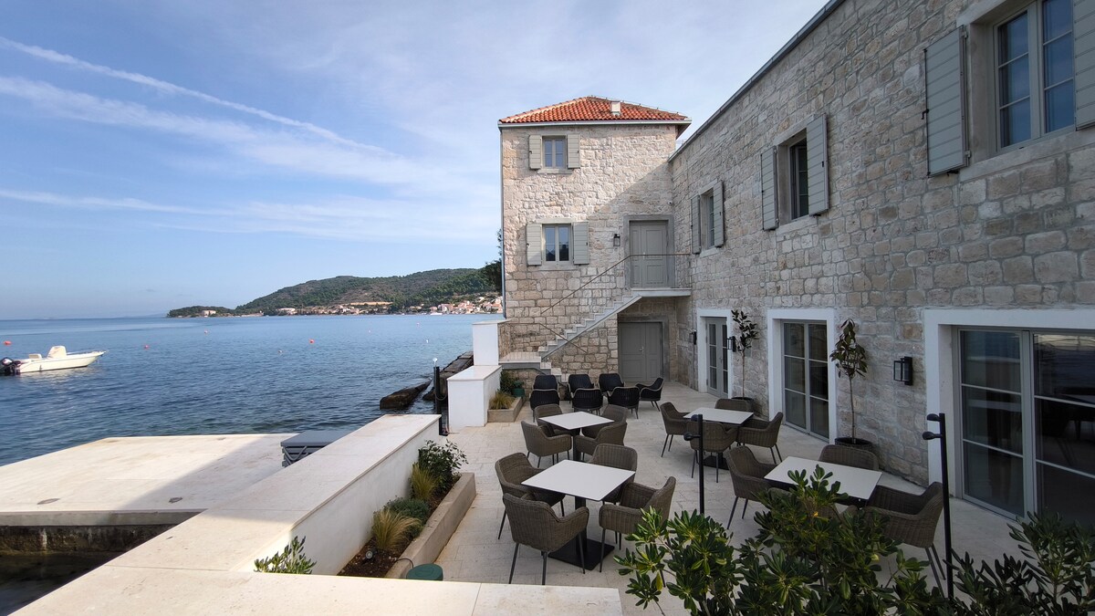 An outdoor terrace offers a serene waterfront view, featuring numerous tables and chairs surrounded by greenery. The stone building is complemented by a clear blue sky and calm water, with boats visible in the distance, creating a peaceful atmosphere.