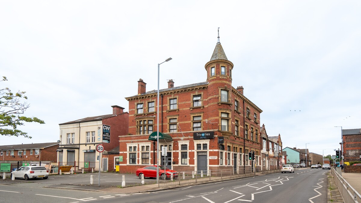 The exterior of the Trivelles Seaforth hotel is depicted, showcasing a red brick building with architectural details. A turret can be seen at the top, and large windows are present on the facade. Cars are parked along the street, with the surrounding area visible.