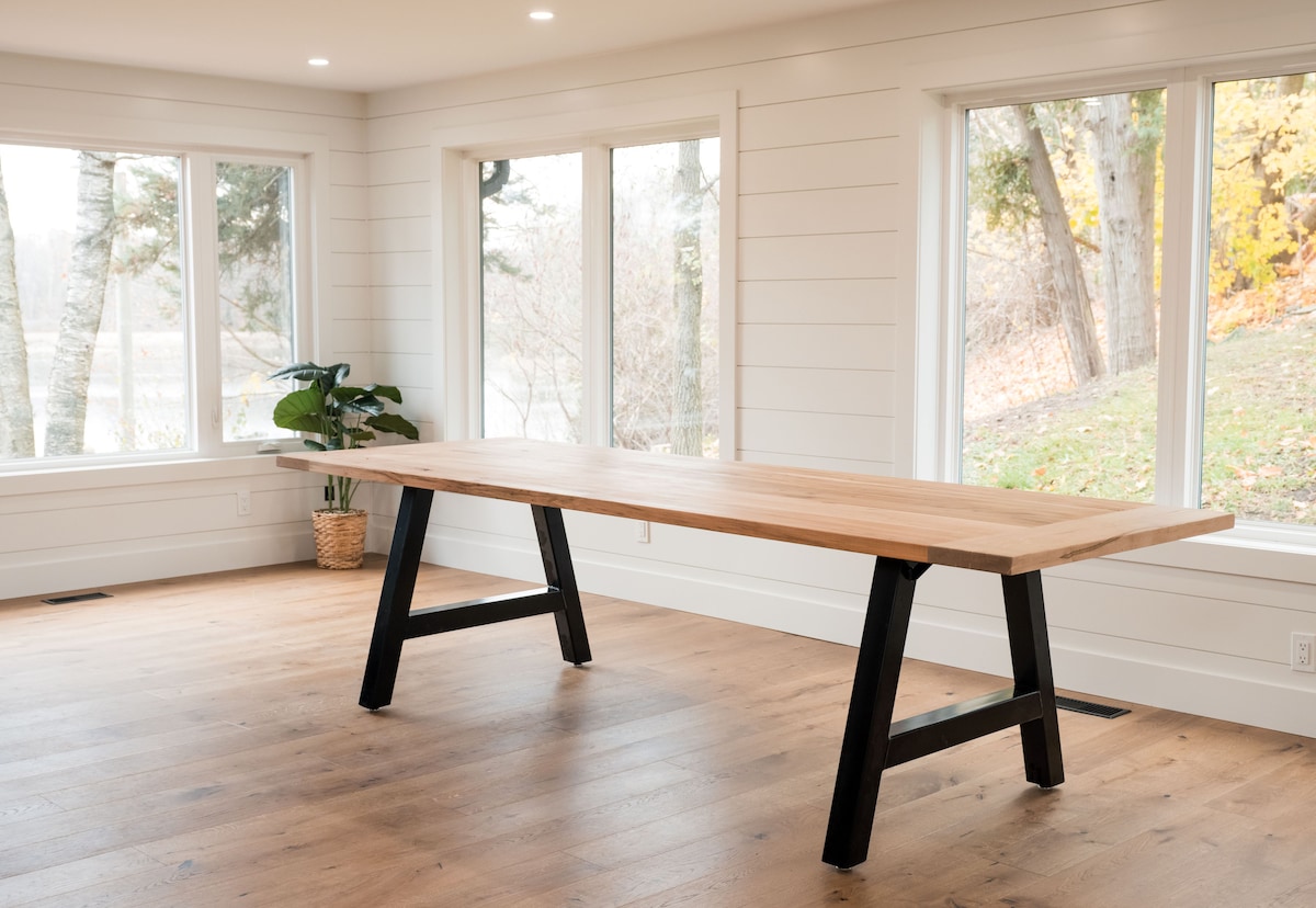 A spacious dining area features a large wooden table with a modern black frame. Natural light floods in through multiple windows, highlighting the fresh greenery of a nearby potted plant and the warm tones of the wooden floor.