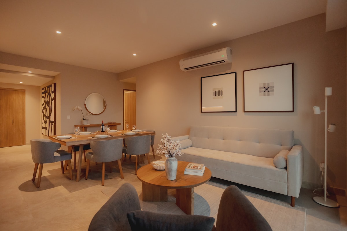 A modern living space is captured, featuring a neutral color palette and comfortable furnishings. A spacious sofa is positioned alongside a circular wooden coffee table. A dining area with a wooden table and accompanying chairs is visible in the background. Artwork adorns the walls, enhancing the decor.