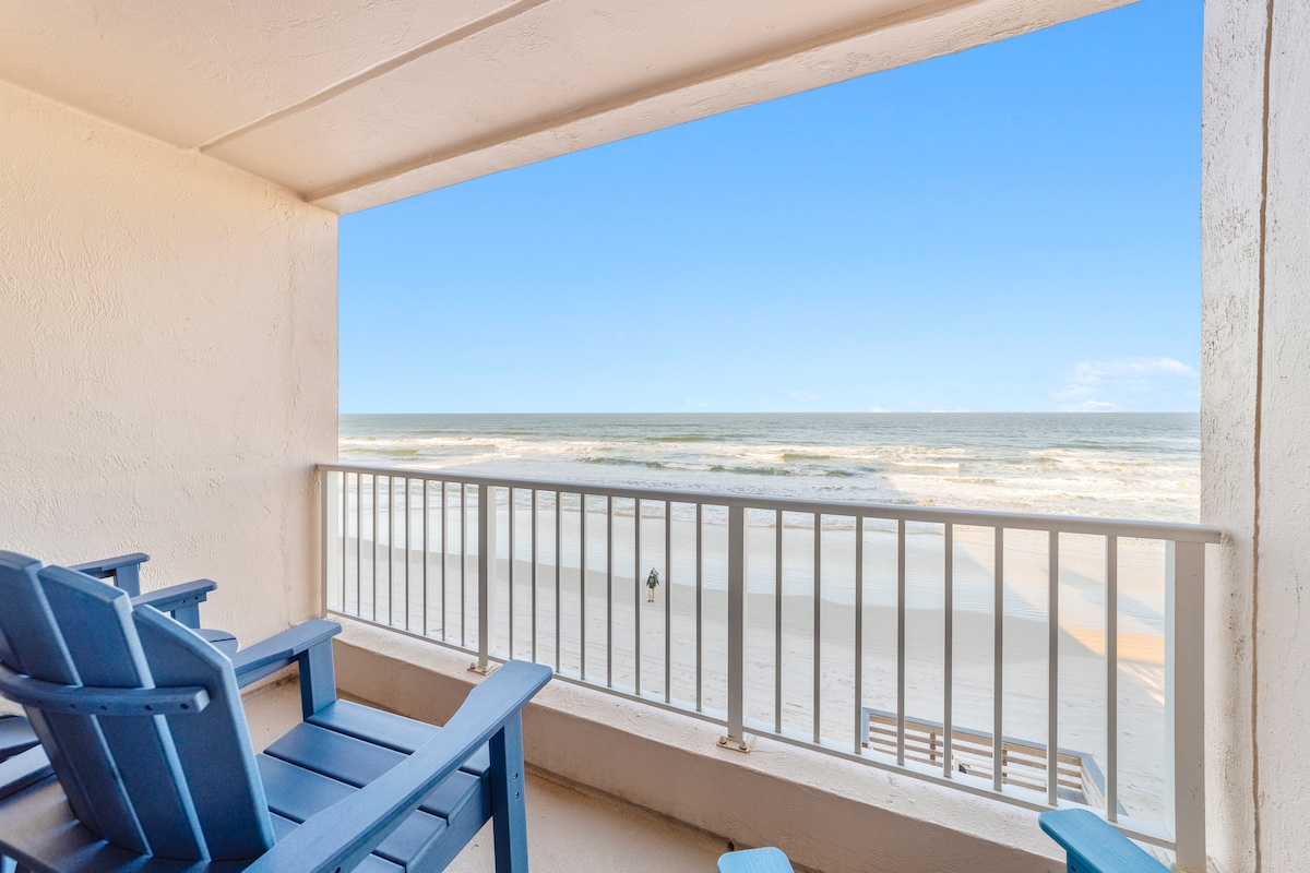 A private balcony overlooks the sandy beach and ocean, featuring several blue outdoor chairs. The panoramic view captures gentle waves lapping the shore under a clear blue sky, inviting relaxation and enjoyment of the coastal scenery.