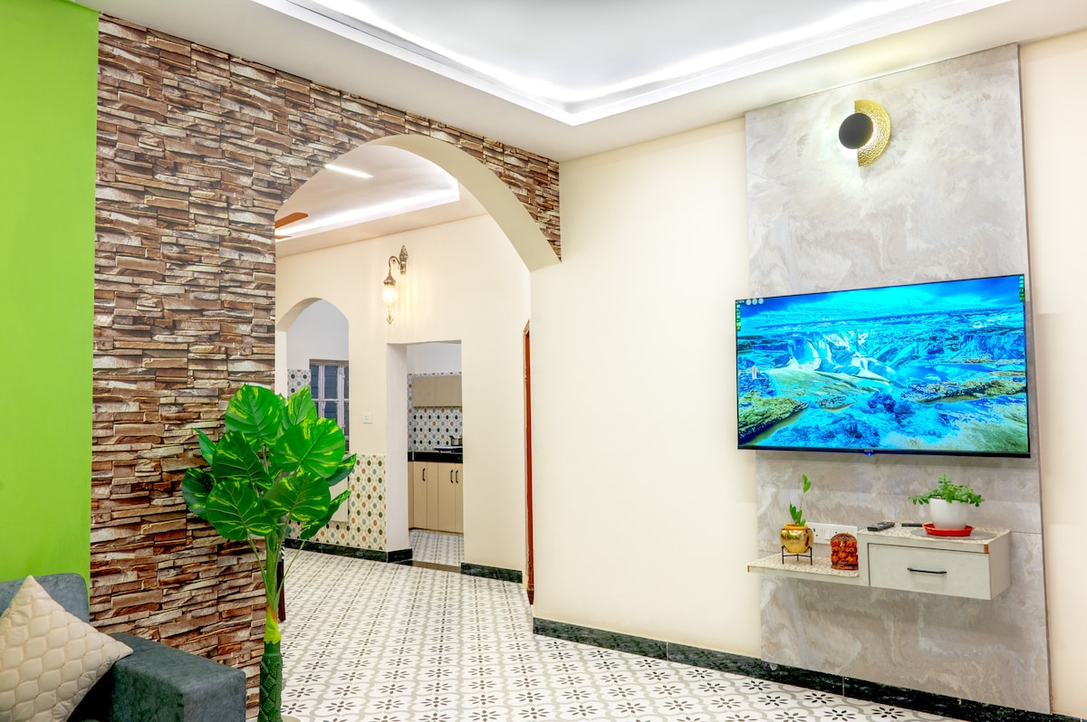 A contemporary living area is showcased with a stylish wall featuring stone accents, complemented by a green accent wall. A modern television is mounted above a minimalist shelf adorned with a small plant. The patterned tile floor enhances the inviting ambiance of the space.