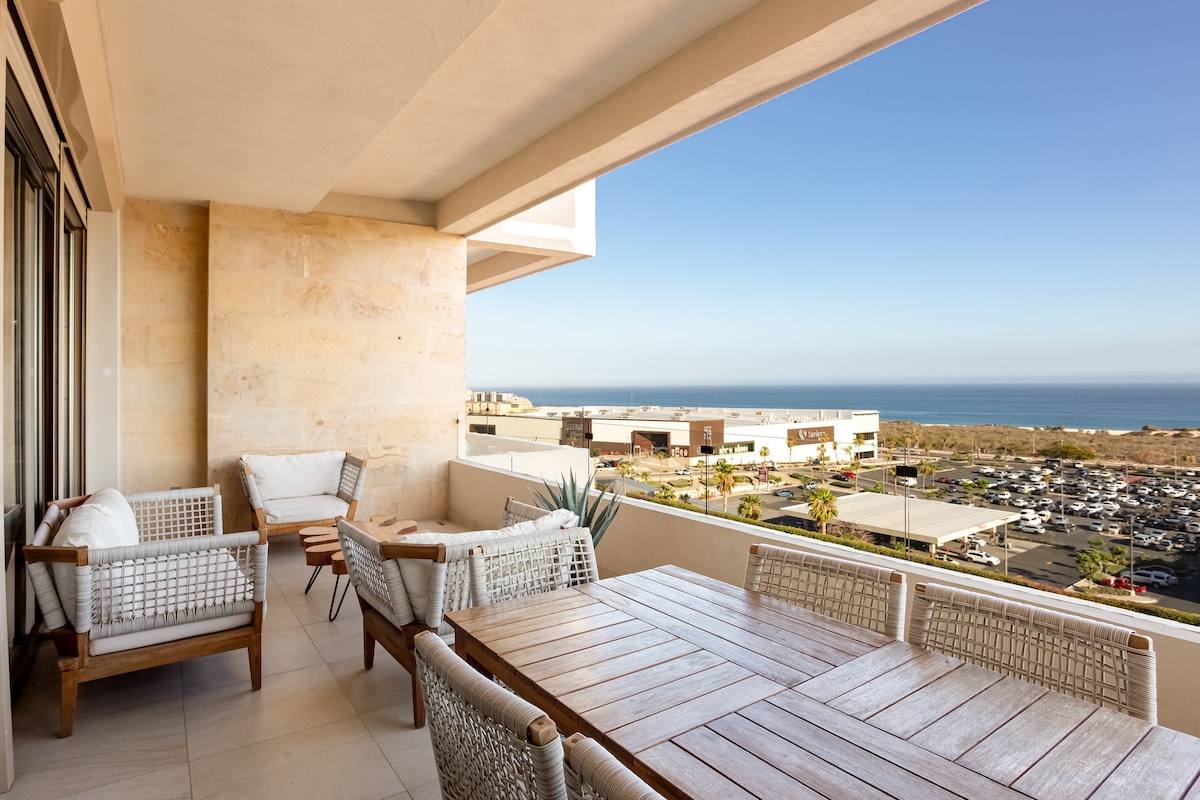 An outdoor balcony is highlighted, featuring a wooden dining table surrounded by comfortable chairs. A gentle ocean view is visible in the distance, complemented by an expansive sky. Natural light brightens the area, enhancing the overall spaciousness.