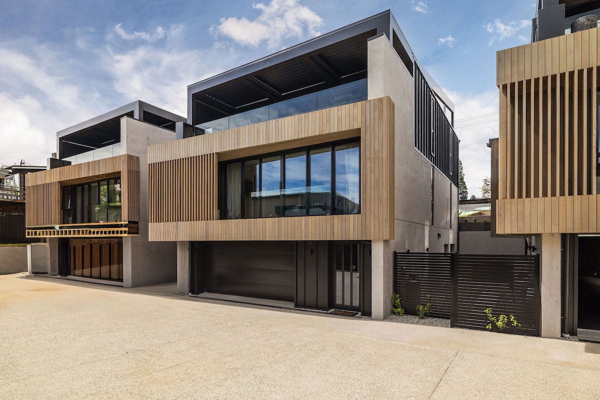 The exterior of the property displays a modern three-level design with a combination of concrete and wooden elements. Large glass windows enhance the facade, allowing natural light to enter. The setting includes a spacious driveway and landscaped areas, contributing to the stylish overall appearance.