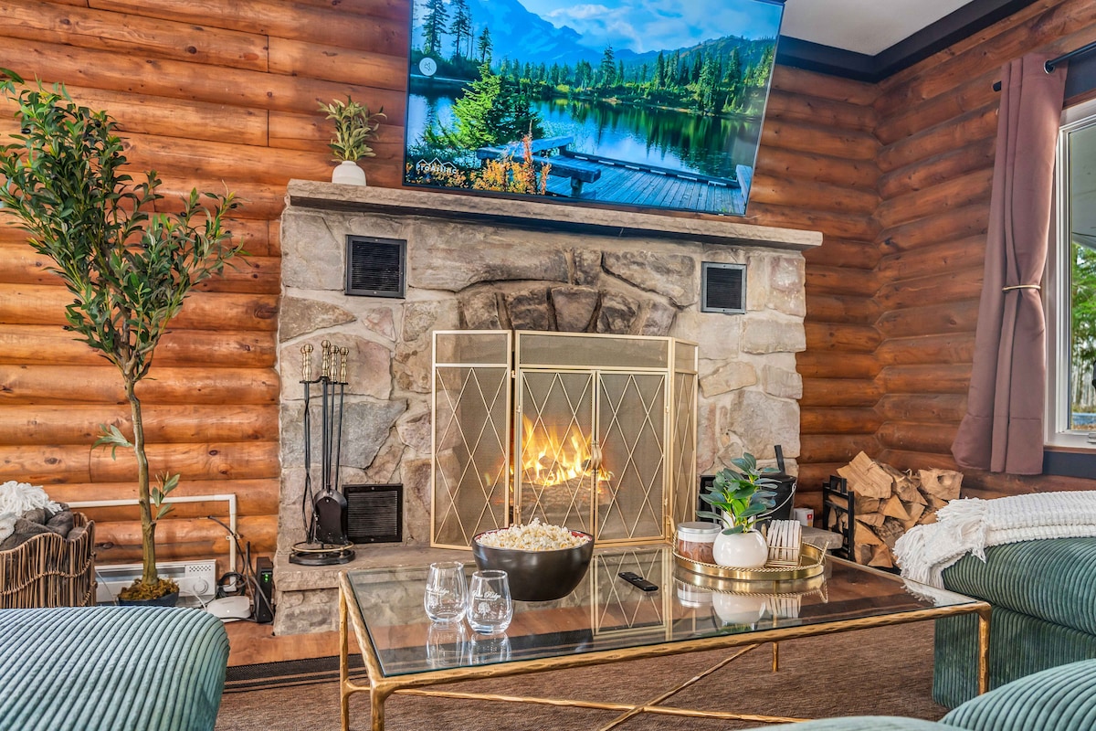 A cozy living area is highlighted by a stone fireplace with a fire gently burning. Above it, a large TV displays a picturesque scene of nature. A glass coffee table sits in front, adorned with decorative items and glassware. Natural wooden walls contribute to the warm ambiance.