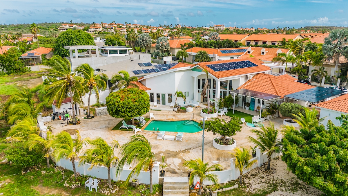 A beautifully landscaped property is presented with a bright turquoise pool surrounded by sun loungers. Lush greenery and palm trees add to the serene outdoor space, while the bright white structure with orange-tiled roofs complements the vibrant coastal scenery.