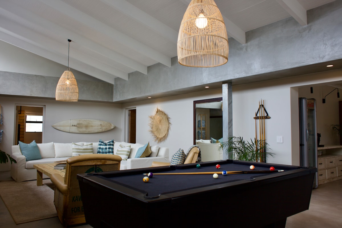 A modern living space features a pool table in the foreground, surrounded by cozy seating and natural textures. Rattan light fixtures hang from the ceiling, illuminating the room. Decorative elements, including surfboards and wall art, enhance the laid-back atmosphere.