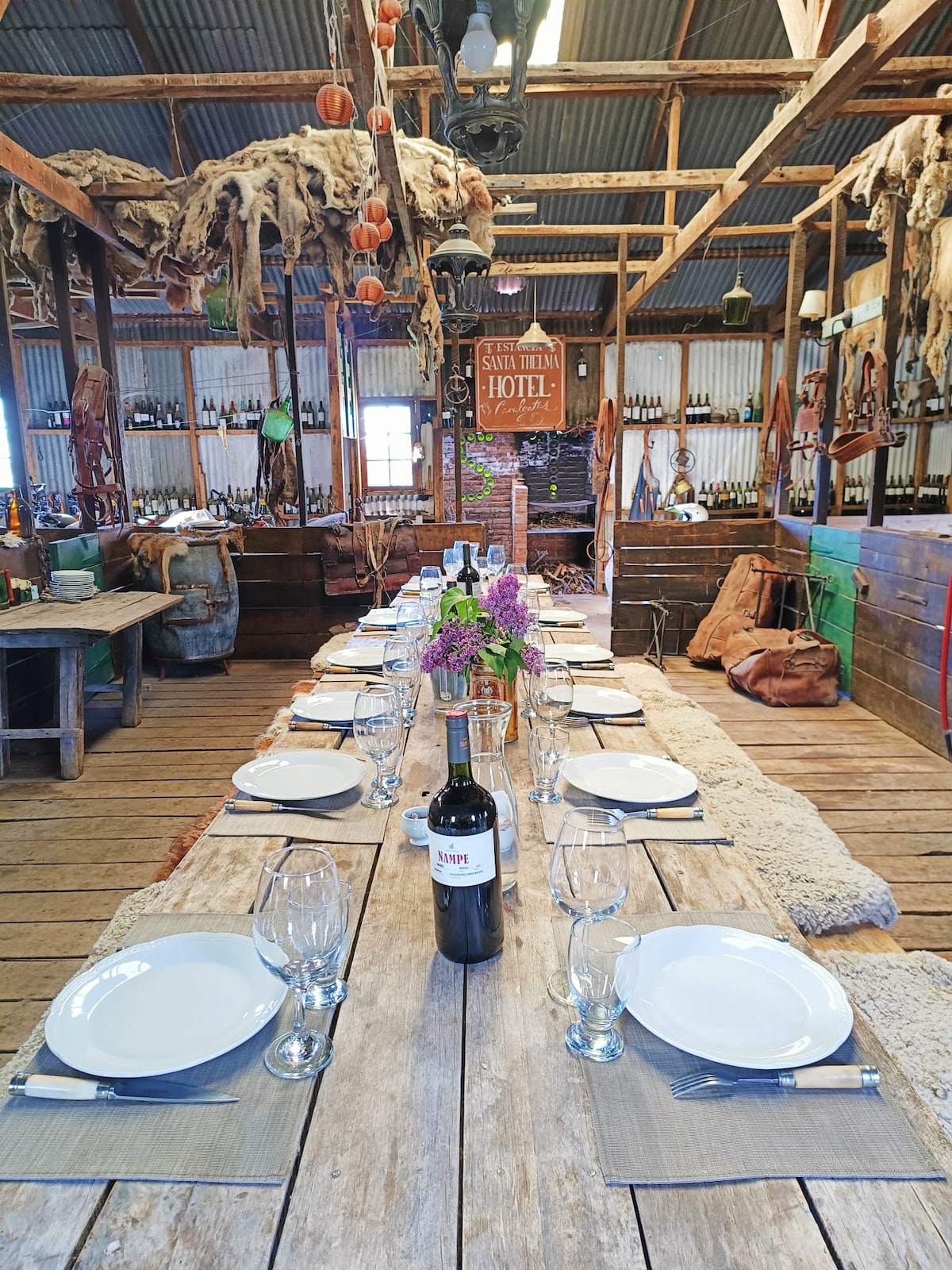 A long wooden dining table is set for guests with white plates, clear glasses, and a bottle of wine at the center. The rustic space features wooden beams, hanging dried herbs, and a cozy atmosphere, complemented by a vibrant floral centerpiece.