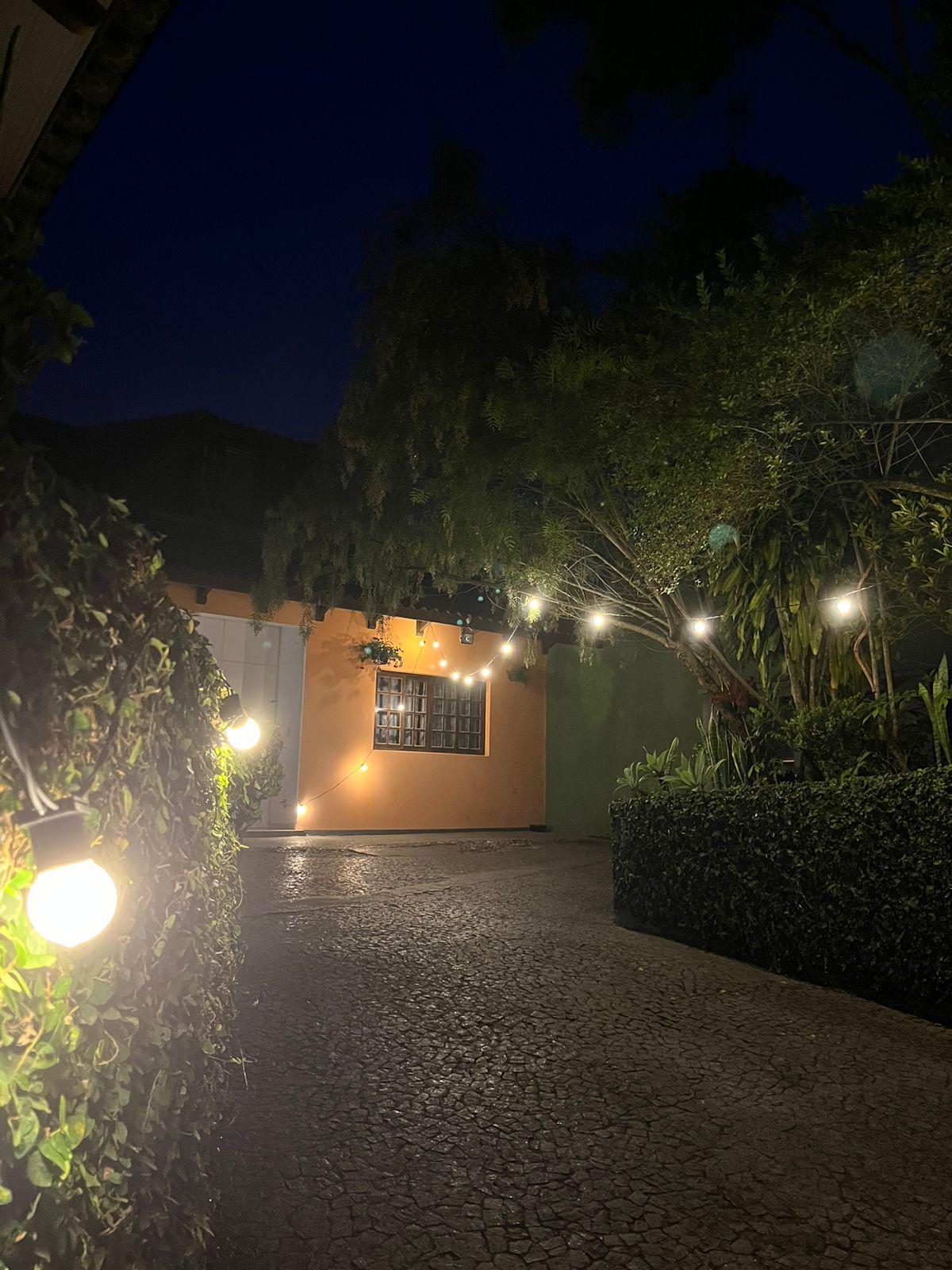 A pathway illuminated by soft overhead lights leads towards a charming entrance. Lush greenery frames the walkway, creating a serene atmosphere. The warm glow contrasts with the dark evening sky, enhancing the inviting feel of the outdoor space.