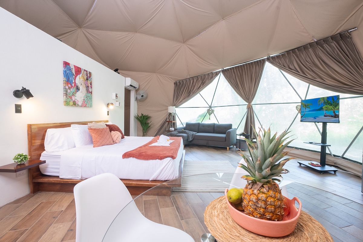 An airy interior of a geodesic dome features a spacious bed with light linens and a decorative throw. A cozy seating area includes a modern sofa and a television. A dining table with a bowl of fruit is positioned in the foreground. Natural light fills the room through large windows.