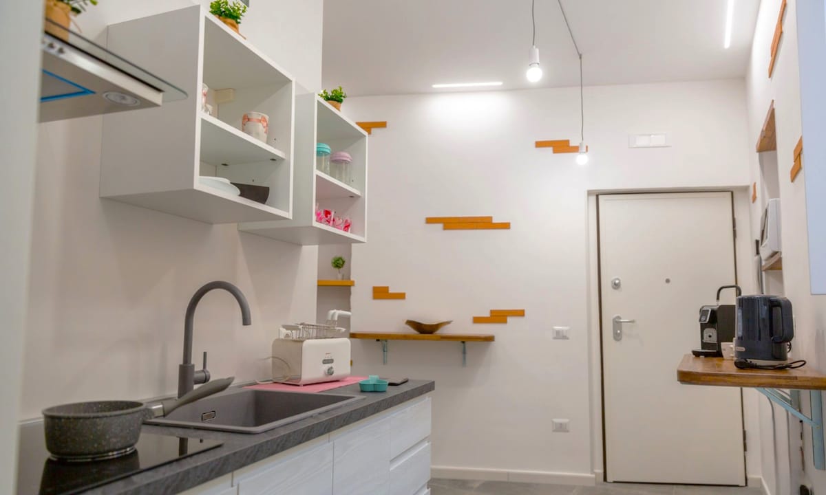 The kitchen area features modern cabinets with open shelving displaying colorful kitchenware. A sleek sink and countertop are complemented by minimalistic decor and warm wooden accents. Bright overhead lighting provides clarity to the space, creating a functional and inviting atmosphere.