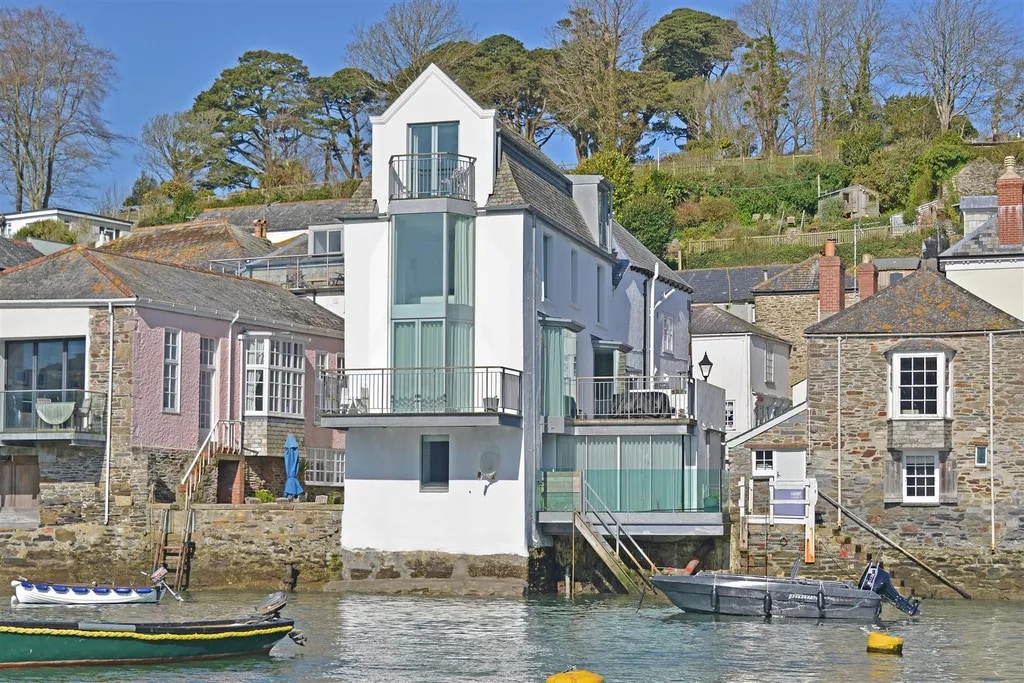A contemporary waterfront townhouse is visible, featuring multiple levels and expansive balconies. The clean lines and large windows provide an open feel. The property is positioned next to the water, with boats nearby, set against a backdrop of lush greenery and coastal homes.
