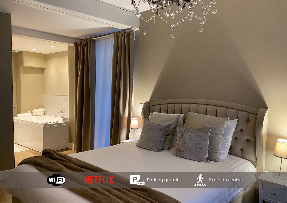 A softly lit bedroom featuring a spacious bed with plush pillows and a stylish headboard. A soft throw covers the foot of the bed. In the background, a luxurious bathtub is positioned invitingly under warm lighting, enhancing the relaxed ambiance of the room.