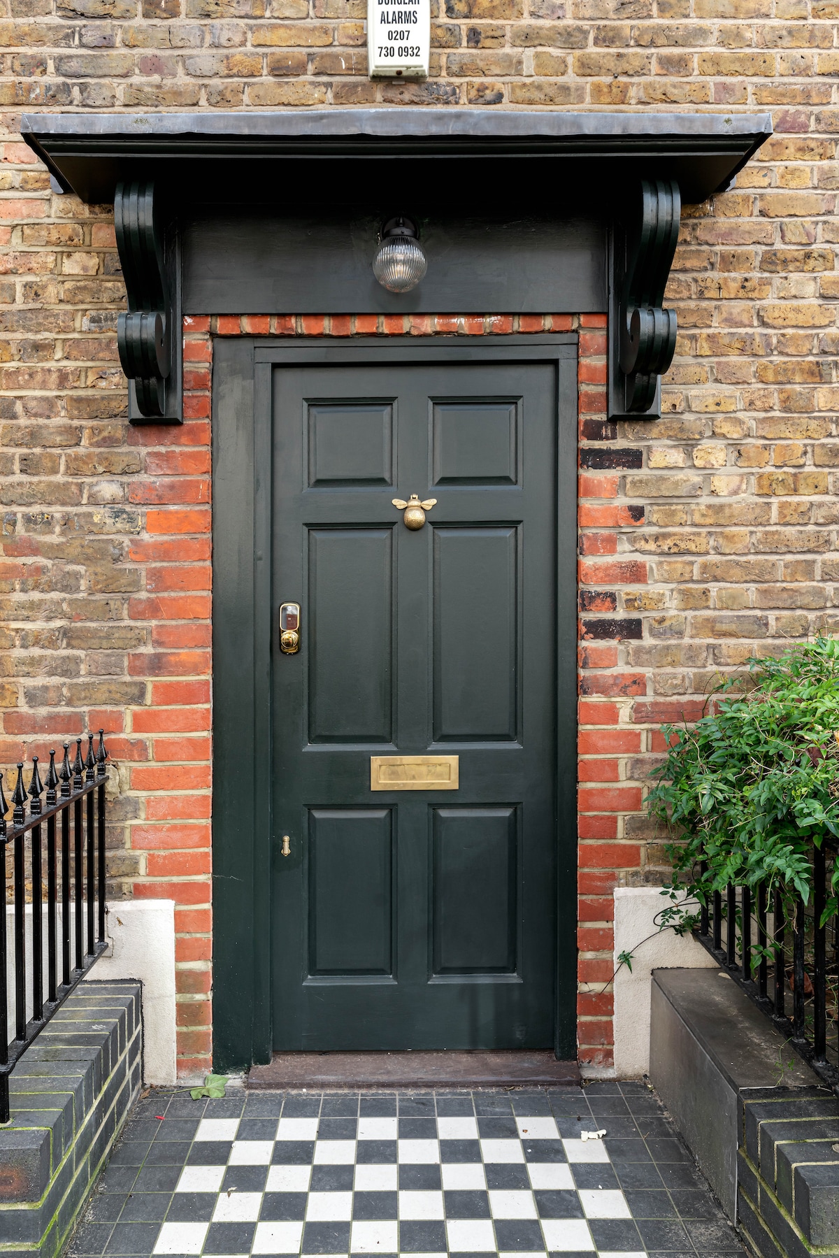 A green door is framed by red brick, featuring a brass door handle and a small brass nameplate. Checkerboard tiles lead up to the entrance, surrounded by a low iron fence and lush green foliage on one side.
