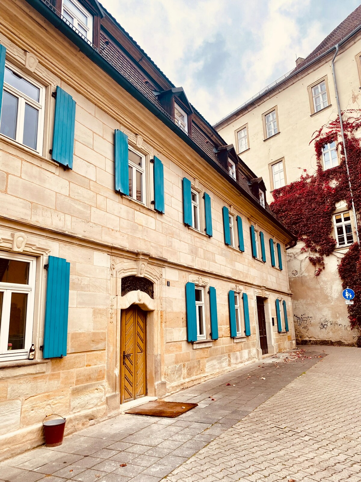 The image features a historical building with a light-colored stone façade complemented by bright blue shutters. An ornate wooden door serves as the entrance, and ivy clings gently to the adjacent walls, adding a touch of natural beauty to the surroundings.