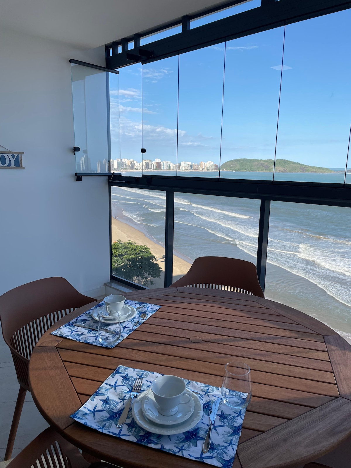 A dining table is set by a large window with a panoramic view of the sea and sandy beach. The round wooden table is accompanied by four chairs. Nautical-themed placemats are laid out, while the azure sky and gentle waves add to the welcoming ambiance.