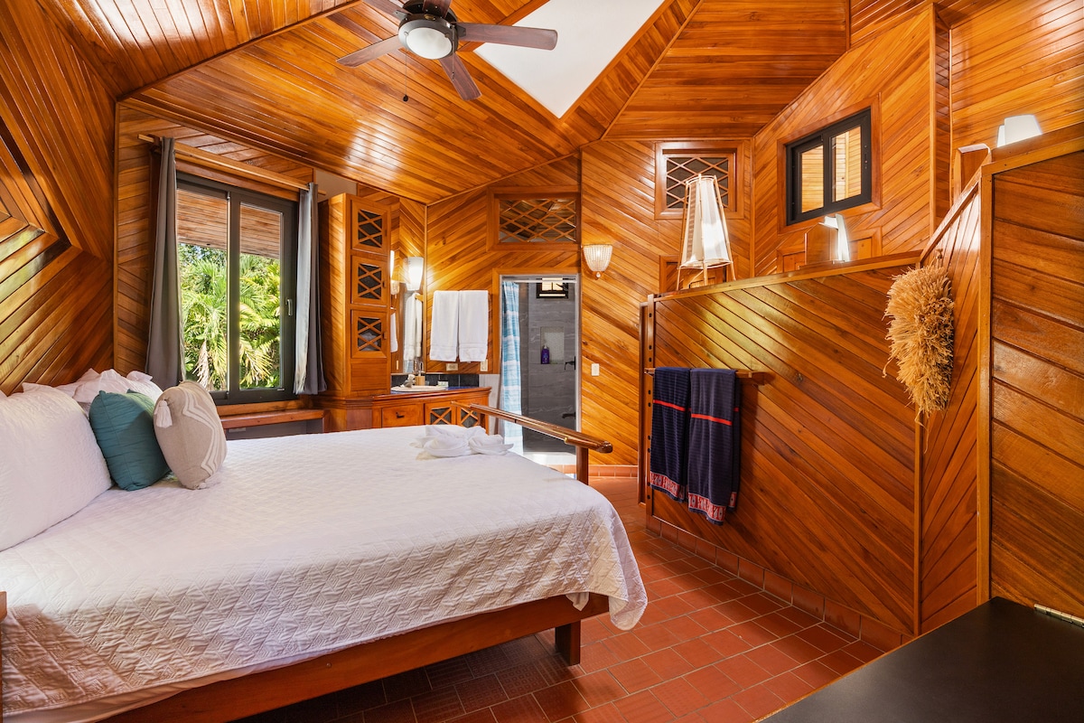 The interior of a bedroom features rich wooden paneling, creating a warm atmosphere. A spacious bed is centered, complemented by decorative pillows. Natural light enters through large windows overlooking lush greenery. A ceiling fan and a subtle light fixture enhance the room's comfort.