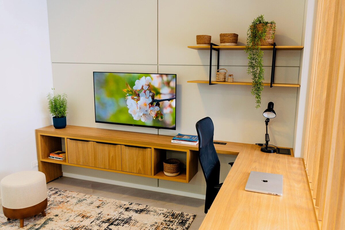 A modern workspace is displayed featuring a light wooden desk with a laptop and a desktop lamp. A large flat-screen TV is positioned above wooden storage, complemented by decorative plants on shelves. A soft ottoman and area rug add comfort to the space.