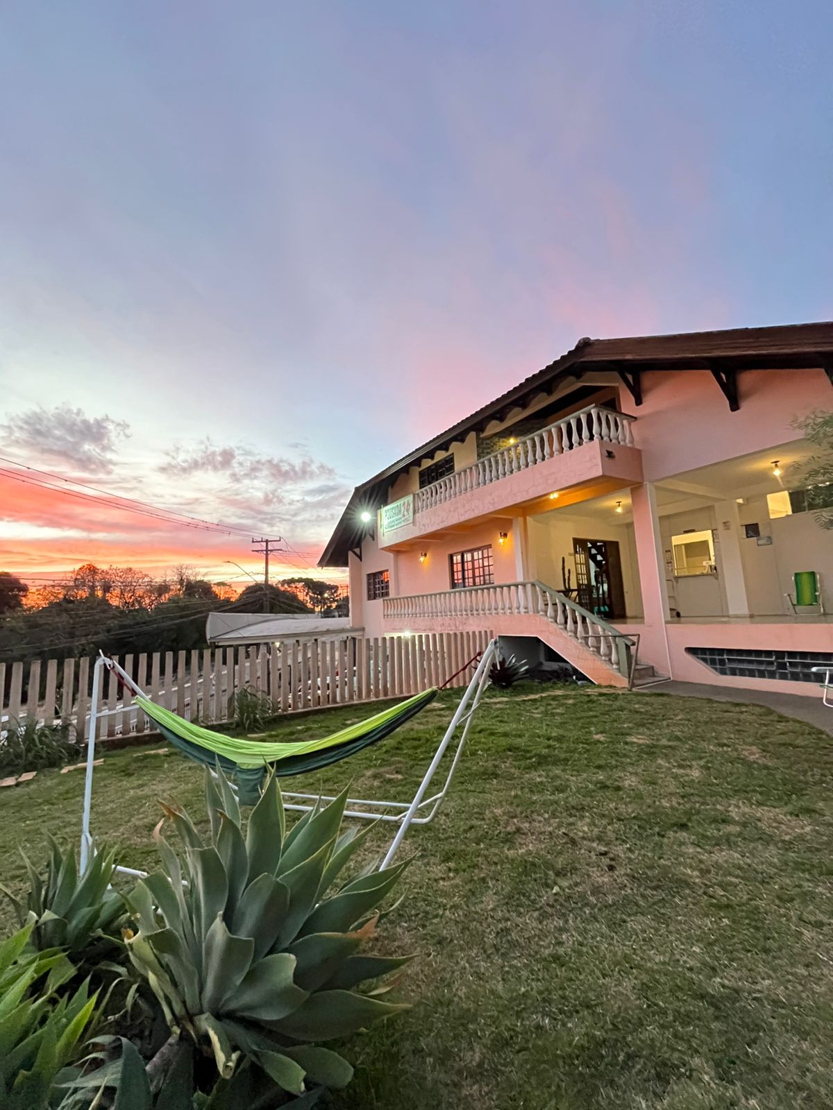 A charming exterior of the hostel is showcased, with a grassy area and a green hammock positioned in front. The building features a welcoming facade with a balcony, and a colorful sunset provides a vibrant backdrop, enhancing the overall ambiance.