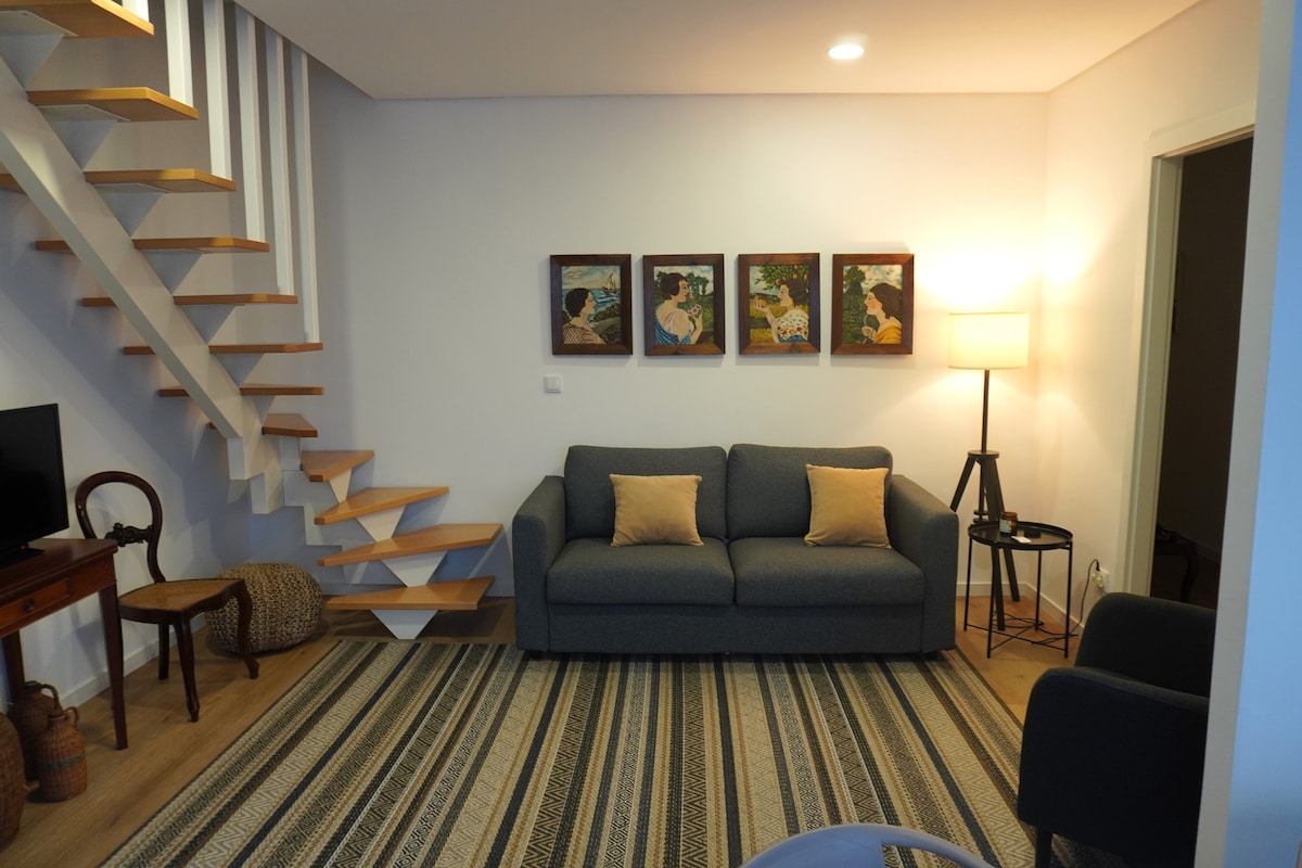 A cozy living area is situated under a staircase with wooden steps. A grey sofa with two yellow cushions faces a wall adorned with three framed artworks. A floor lamp stands beside a small table, and a patterned area rug spans the hardwood floor.
