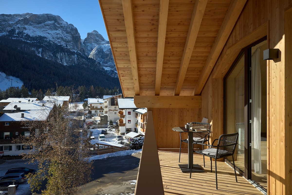 A spacious balcony features a wooden floor and is furnished with a small table and two chairs. The surrounding mountains provide a scenic backdrop, while the view encompasses nearby buildings and snow-covered areas.