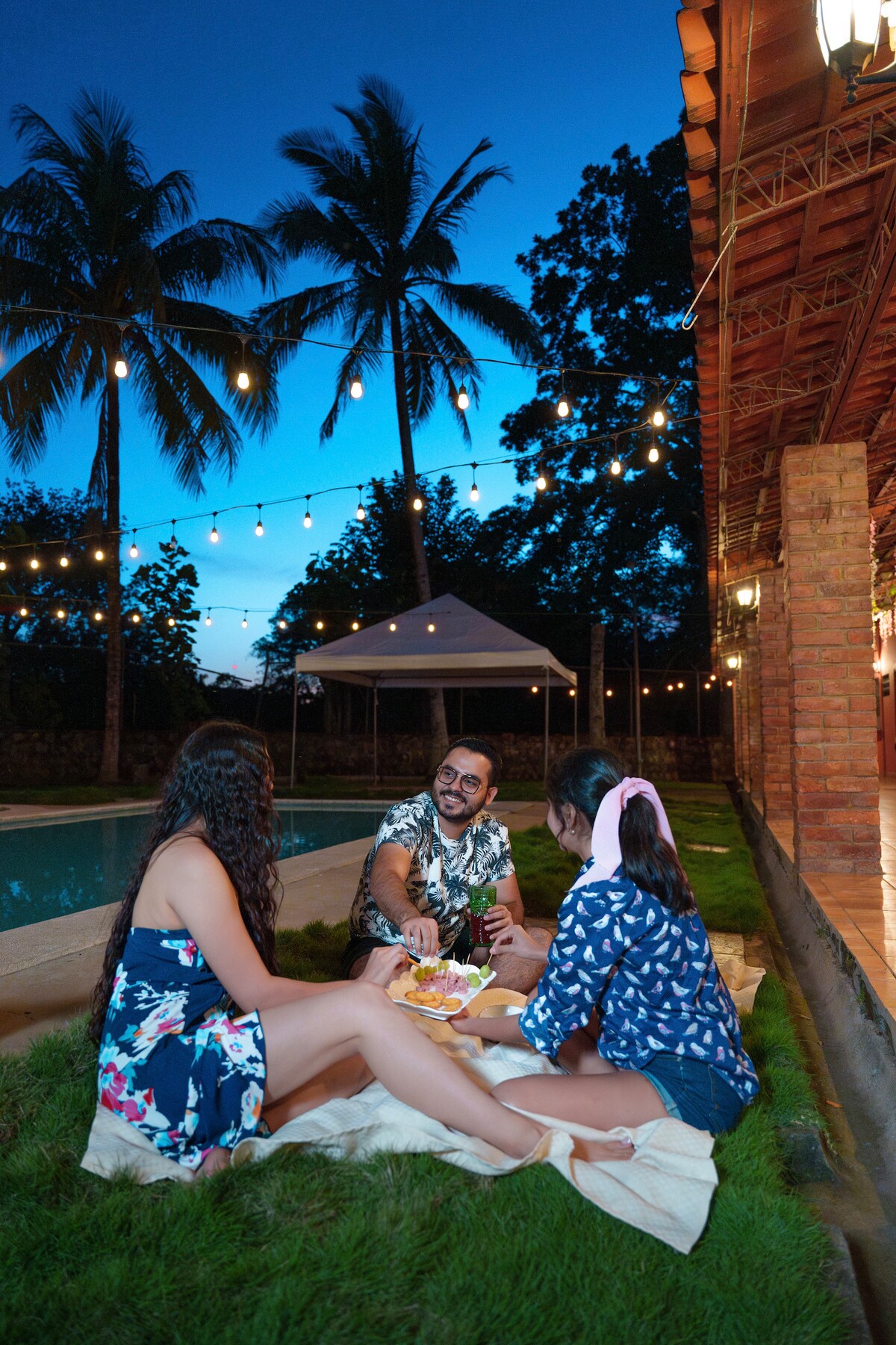 A serene outdoor setting features guests gathered on a blanket under string lights, with a pool visible in the background. Lush green grass surrounds the area, bordered by palm trees, while a tent provides shaded seating for a relaxed dining experience.