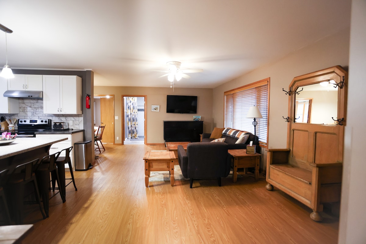 An open living area features a comfortable seating arrangement with a dark armchair and light wooden coffee tables. A large TV is mounted on the wall, and bright natural light enters through the windows. The kitchen area is visible in the background, showcasing modern appliances.