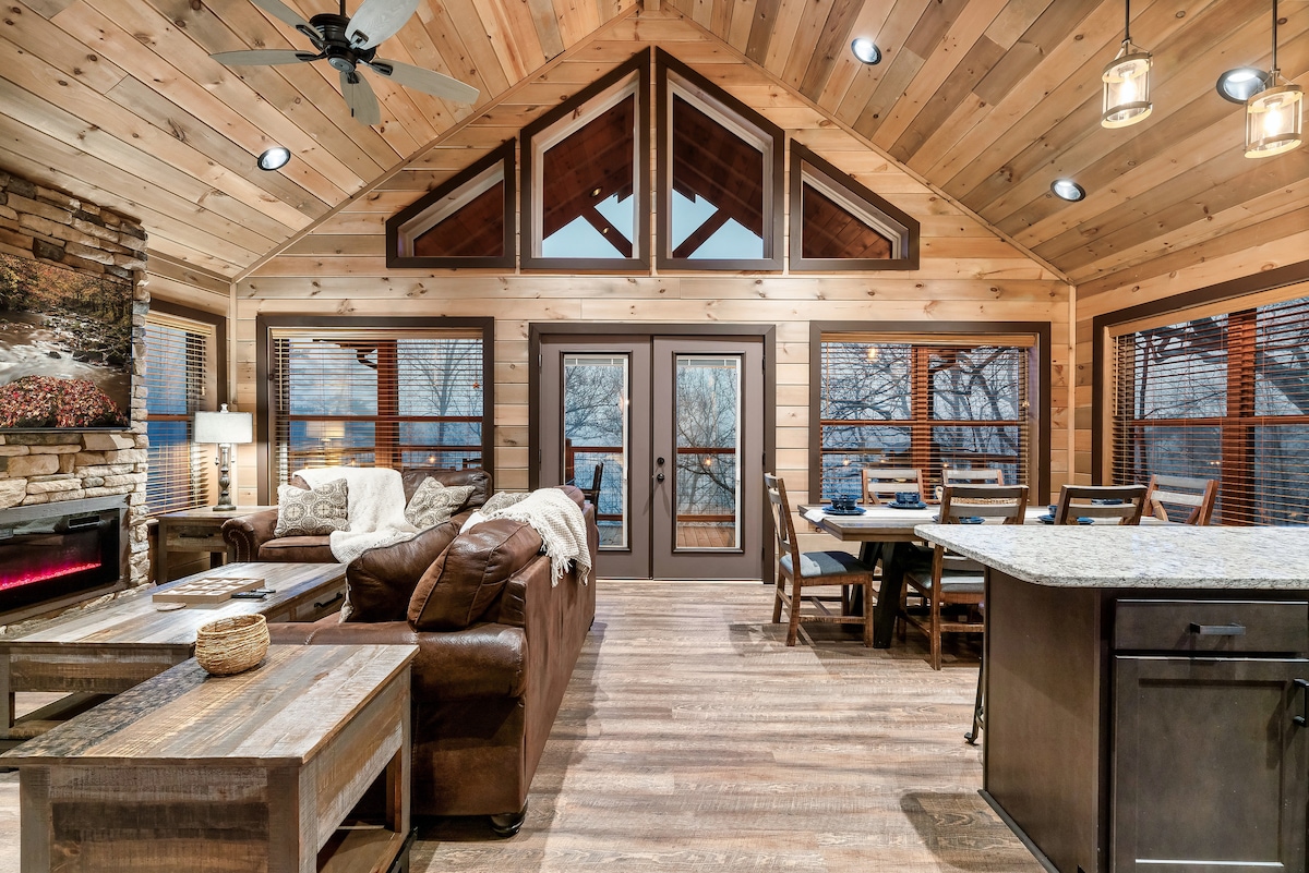 The spacious living area features vaulted ceilings with wooden beams and large windows overlooking the outdoors. A comfortable brown sofa is positioned near a stone fireplace, with a rustic coffee table in front. A dining table set for six is visible in the background, adding to the inviting atmosphere.
