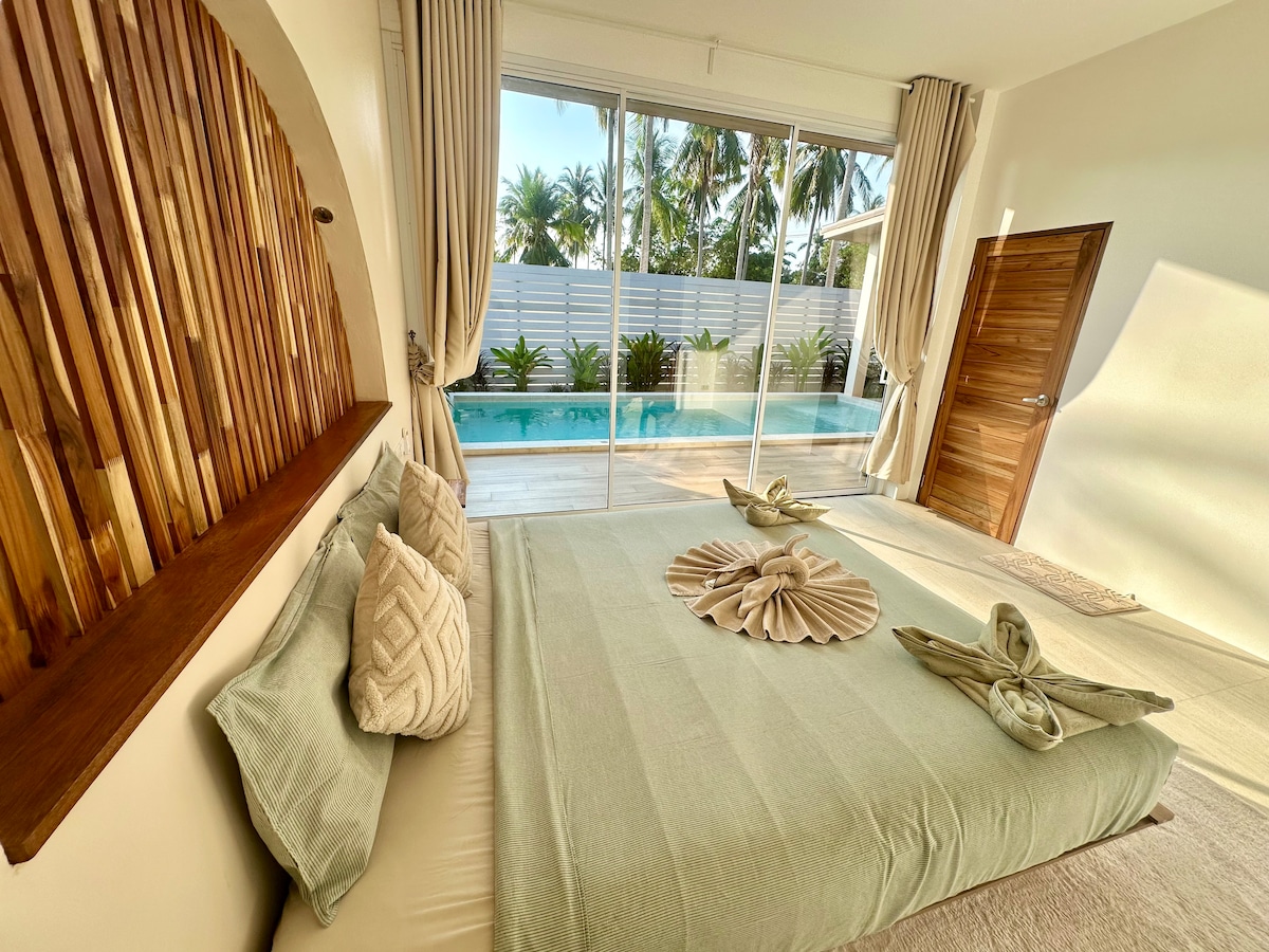 A spacious bedroom features floor-to-ceiling windows that provide a view of the pool area surrounded by tropical plants. Soft, muted tones of the bedding create a calm environment, while decorative folded towels are elegantly arranged on the bed.
