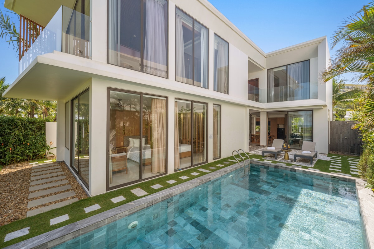 A modern villa exterior showcases large glass doors and a private pool. The pool is surrounded by a stone patio with lounge chairs, providing a tranquil space for relaxation. Lush greenery frames the property, enhancing the serene atmosphere under clear blue skies.