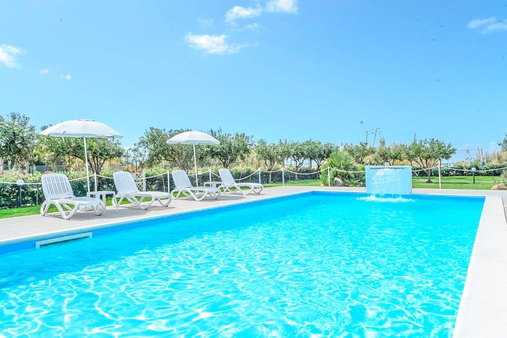 A sparkling blue pool is surrounded by several white lounge chairs and umbrellas, providing shade. Lush greenery and trees are visible in the background, contributing to a serene outdoor atmosphere.