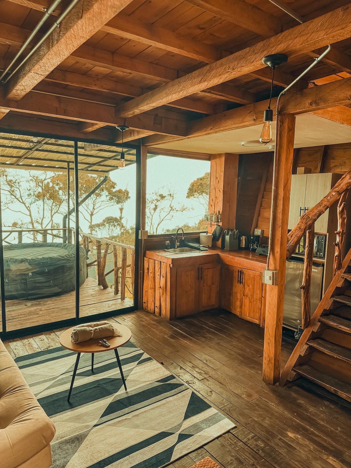 An open-concept interior showcases rich wooden beams and a cozy lounge area with a beige sofa centered on a patterned rug. A small table offers a space for relaxation, while a rustic kitchen area features wooden cabinetry. Large glass doors lead to outdoor scenery.
