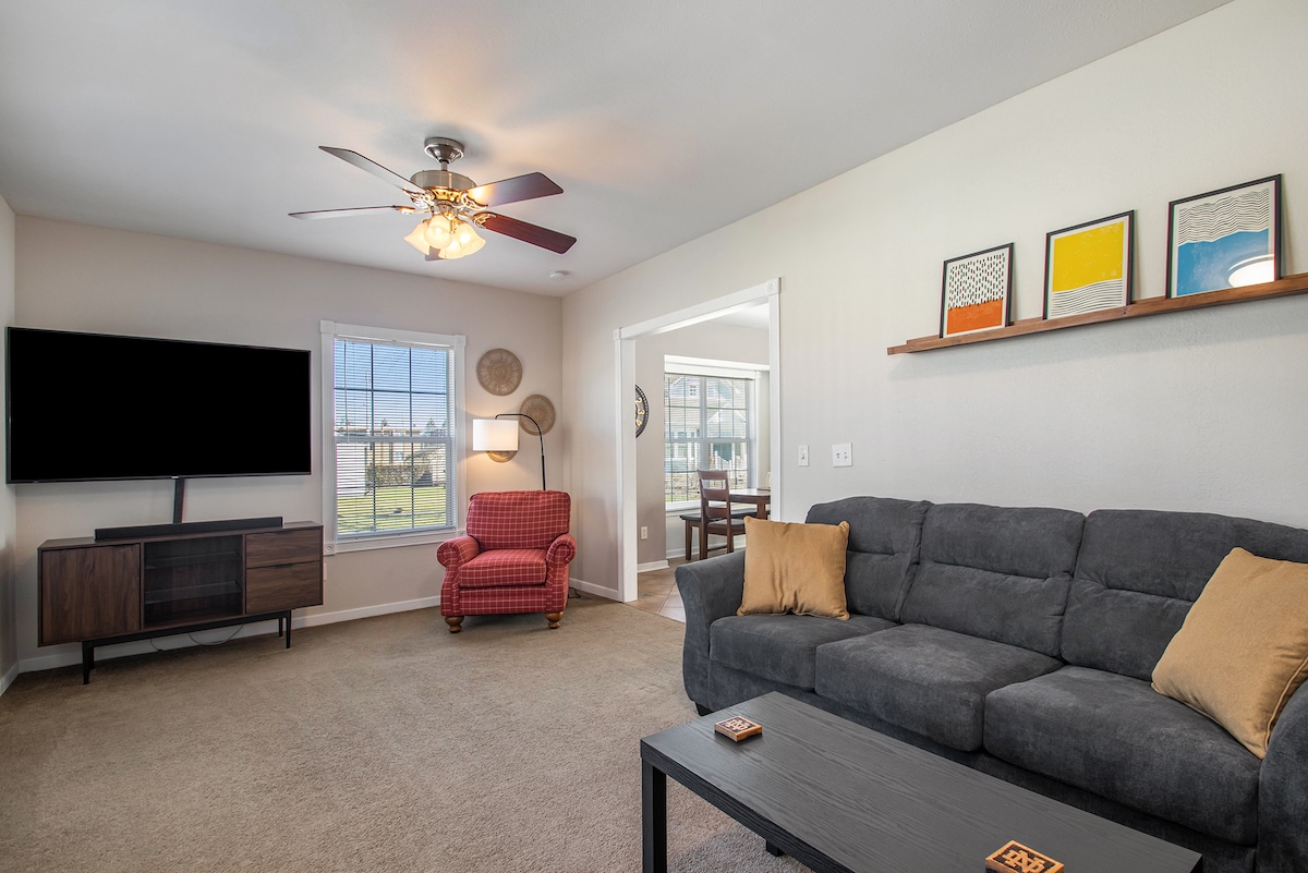 A spacious living area is furnished with a dark gray sofa and an armchair in a warm red hue. A 70-inch TV is mounted on the wall opposite the sofa. Natural light fills the room through large windows, highlighting the soft carpeted flooring.