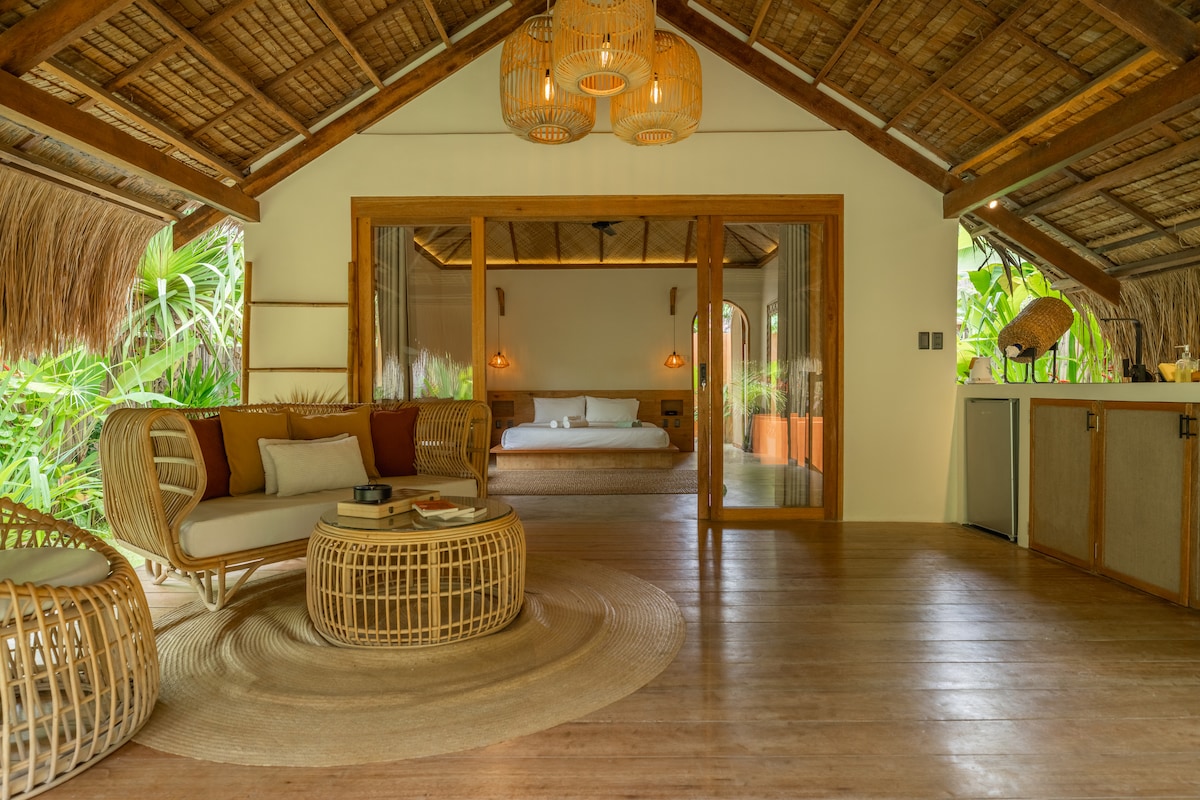 An airy living area showcases natural materials and design, featuring a spacious seating arrangement with cozy cushions. Light pours in through large glass doors, leading to an inviting bedroom area, while greenery peeks through the windows, enhancing the indoor-outdoor experience.