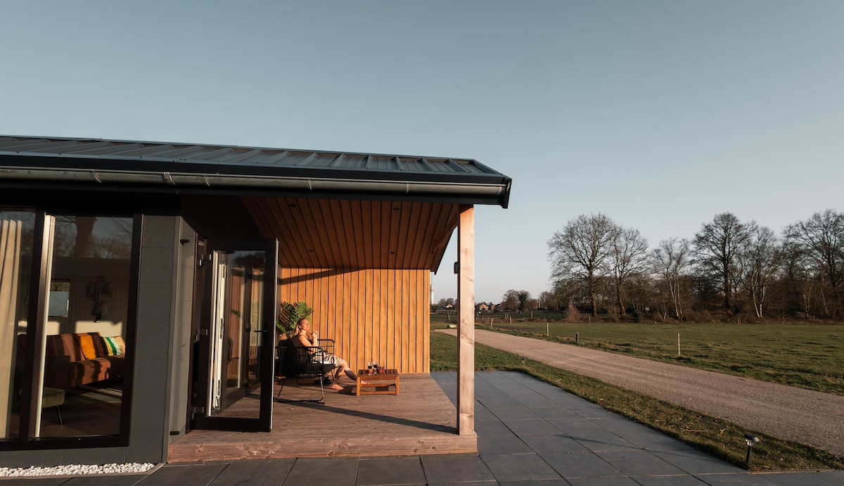 A modern chalet features a covered wooden porch, offering comfortable seating for relaxation. Large glass doors provide an inviting entry to the interior. The surrounding landscape showcases an open field and a gravel path, bordered by trees against a clear sky.