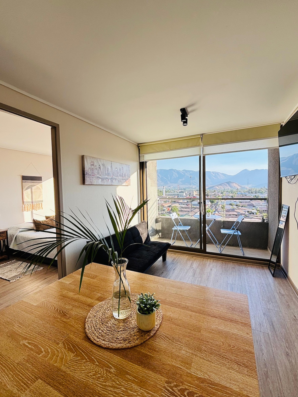 A modern living space features a wooden table with a small potted plant, complemented by a black couch. Large glass doors provide a view of the surrounding mountains and cityscape, while two balcony chairs are positioned outside, enhancing openness and connectivity to the outdoor scenery.