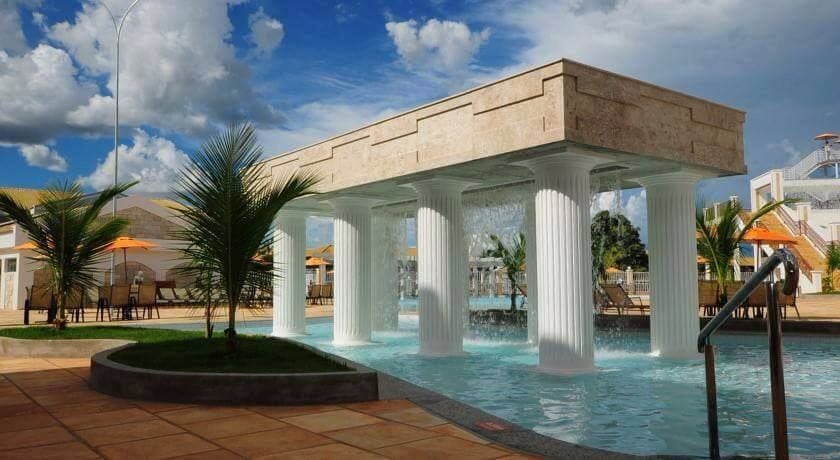 A serene water feature is framed by columns, creating an elegant ambiance. Sun loungers and umbrellas are visible in the background, surrounded by lush greenery and a clear sky. The vibrant blue water enhances the inviting atmosphere of the outdoor space.