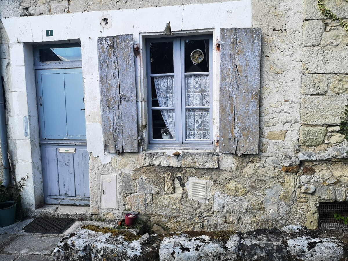 A rustic stone wall features a blue window with shutters, showcasing lace curtains behind the glass. Next to the window, a weathered door with a matching color adds character. Small decorative elements, such as a planter, are positioned on the ground, enhancing the inviting exterior.