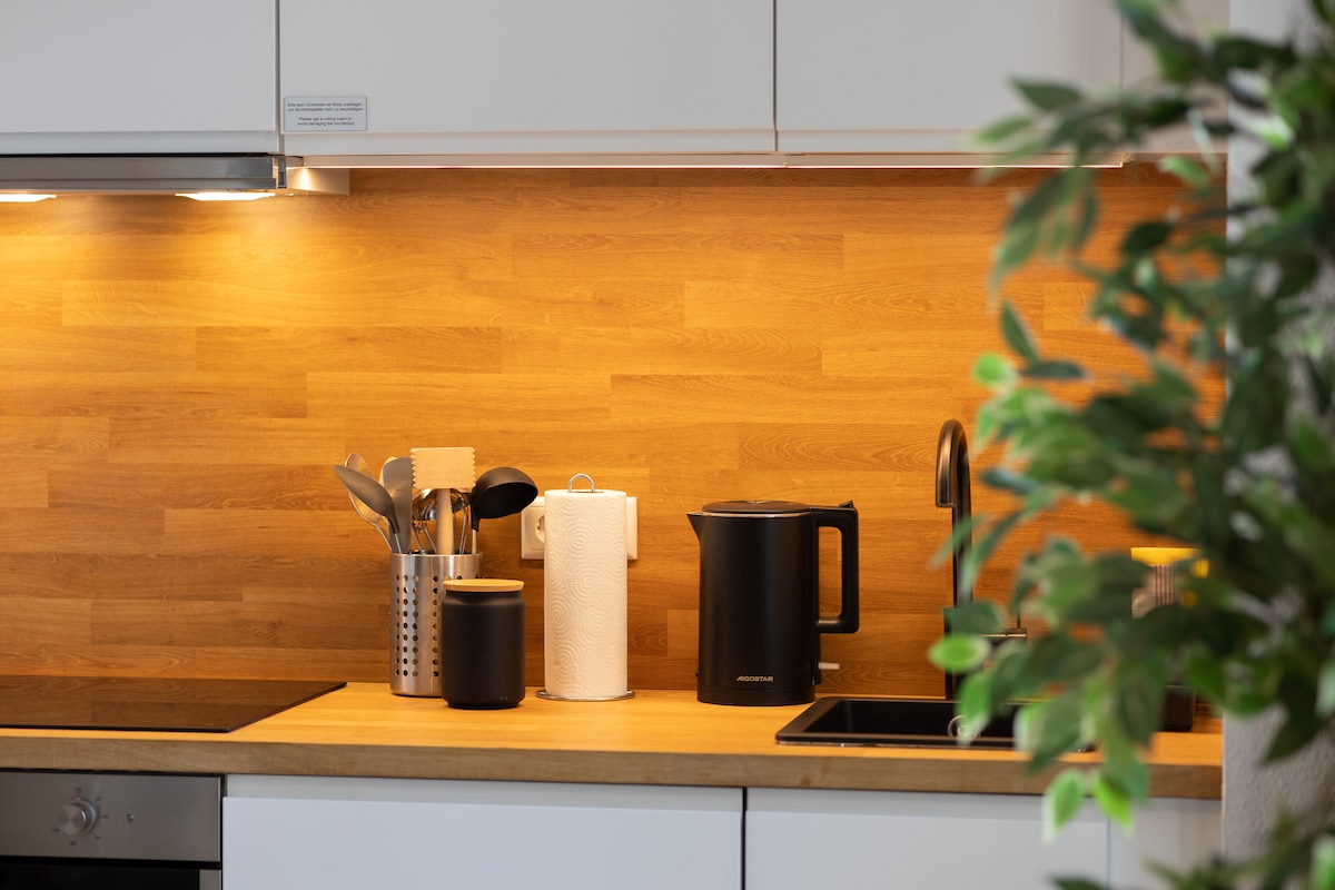 A modern kitchen displays warm wood accents and sleek cabinetry. Essential kitchenware is neatly arranged, including utensils, a paper towel holder, and an electric kettle. A potted plant softly frames the scene, adding a touch of greenery to the functional workspace.
