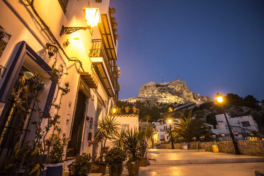 The charming street is lined with traditional white buildings, featuring wrought iron balconies and illuminated by soft yellow lights. Lush green plants add a natural touch, while the imposing mountain with a castle silhouette rises majestically in the background, creating a serene evening atmosphere.