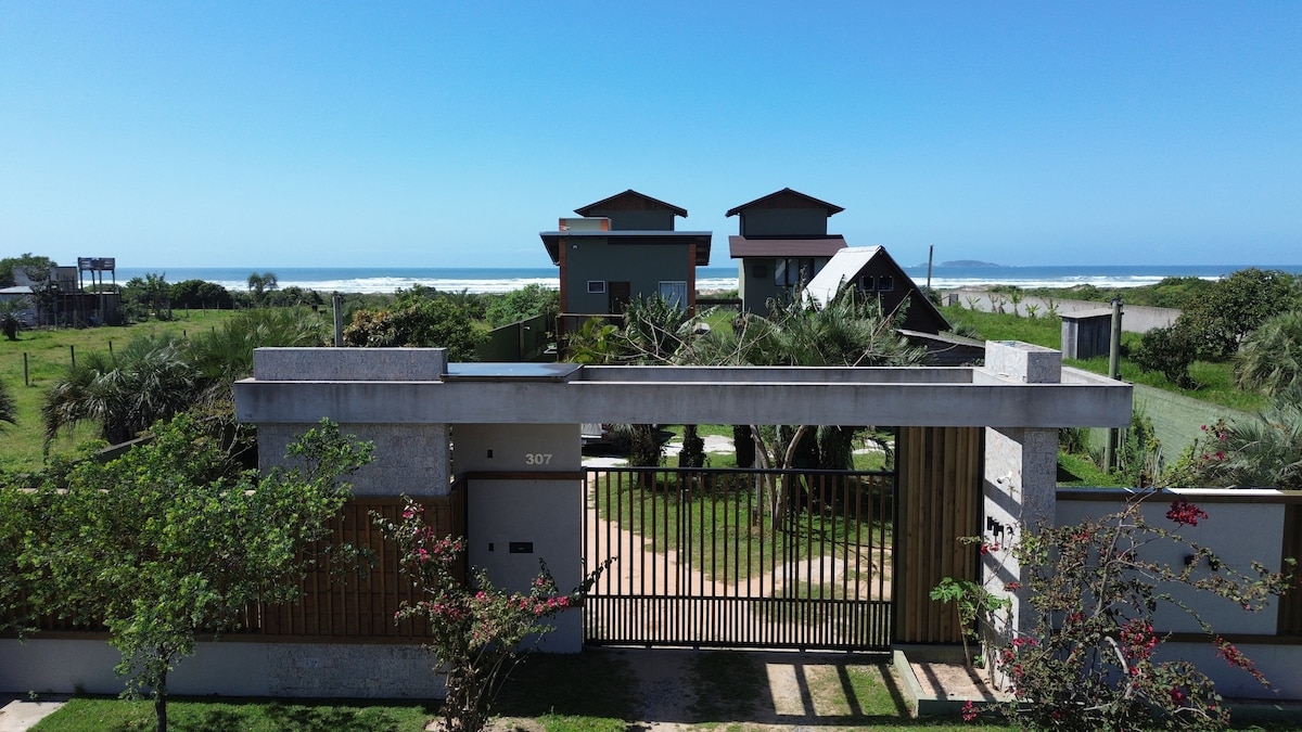 A welcoming property entrance is visible, featuring a modern gate leading into a landscaped garden. Beyond the gate, two towers of apartments are situated, surrounded by greenery and with the ocean visible in the background, creating a serene coastal atmosphere.