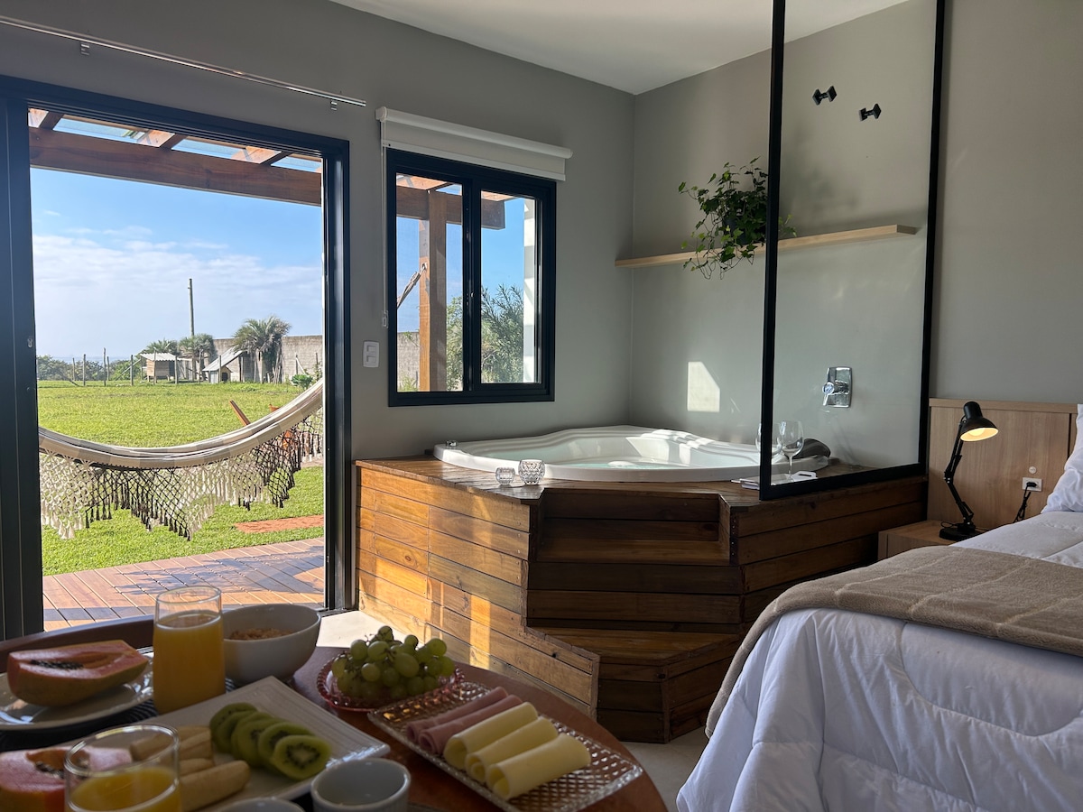 The image displays a modern space featuring a spacious bed, a wooden jacuzzi prominently placed beside a large glass window. A hammock is visible outside, providing a relaxing view of the garden. A breakfast setup with fruits and beverages is arranged on a table.