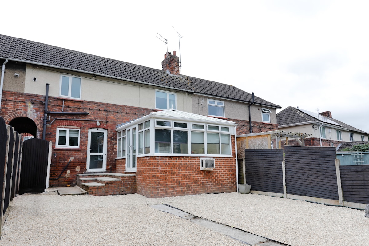 The rear view of the house showcases a spacious outdoor area with gravel flooring. A conservatory extends from the main structure, providing additional light and space. The property is enclosed by wooden fencing, ensuring privacy in the backyard.